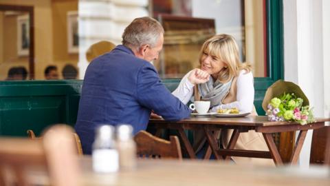 A man and a woman sat opposite each other at a table. They have coffee and plates of food and there is a bunch of flowers on the table next to the woman. They are mid conversation, smiling at each other. 