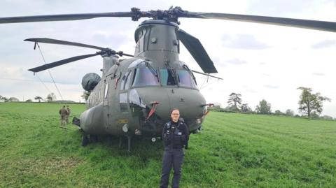 A police officer is standing in front of a Chinook helicopter which is stationery in the middle of a field. 