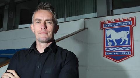 Kieran McKenna wearing a black shirt standing next to the Ipswich Town Football Club crest. He is looking directly into the camera and has his arms crossed.