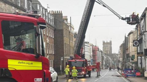 High West Street in Dorchester with two fire engines, one with an aerial platform with two firefighters raised to roof level along one side of the street