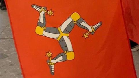 A close-up of three Manx flags, which are red with a yellow, white and grey three-legs of man symbol. 