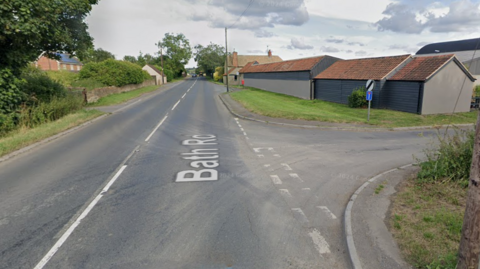 The junction at the A365 and Bowerhill lane. There are farm-like buildings to the right of the carriageway and a grassy verge either side.