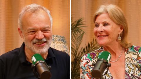 A man with white hair and a woman with shoulder length hair speak into microphones. The woman is wearing a patterned blouse.