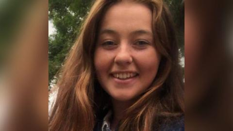 A picture of a young woman with auburn hair smiling. A tree is in the background.
