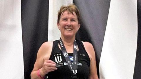 Carole Munro smiles at the camera wearing a medal and holding a patch. She is standing in front of a black and white striped backdrop and is wearing a black vest top.