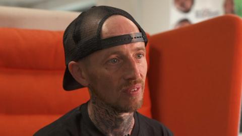 Steven, a man in a black t shirt and black snapback cap sat on a large orange chair. He has a short red haired goatee.