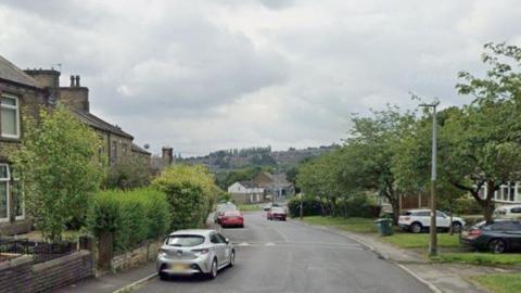 A residential street with terraced houses, trees and parked cars on both sides of the road. 