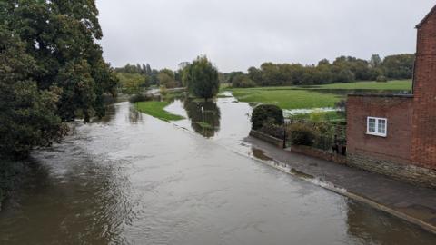 A river can be seen on the left-hand side of the picture which has spilled out onto a path and a field. A house can also be seen in the front-right of the image. A fairly large part of the field has flooded with trees in the background and some overhanging the river