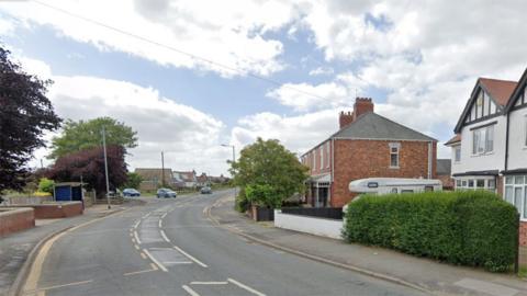 Google Street View looking towards Queen Street from Hollym Road in Withernsea with cars coming out of a junction. There are terraced houses to the right, with a garden hedge at the front of one house and a motorhome parked in the driveway of another.