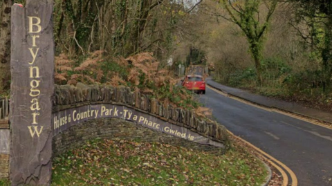 A tree-lined country road curves gently through a wooded landscape at the entrance to Bryngarw House and Country Park. In the foreground, a tall vertical stone marker carved with the word “Bryngarw” stands beside a curved stone wall bearing bilingual Welsh and English signage for the park. Brown ferns, ivy-covered trees and fallen leaves surround the entrance. A small red car is visible further down the road, which disappears into the wooded park beneath a canopy of bare branches.