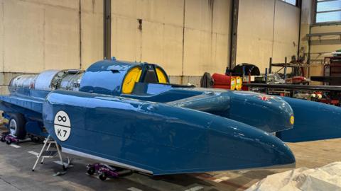 Bluebird, which is a blue hydroplane sitting in a workshop. It looks similar to a fighter jet plane but does not have wings.