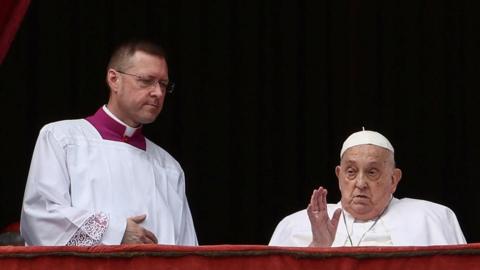 Pope Francis sits and waves from a chair on a balcony next to a colleague.
