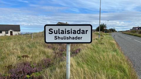 Dealbh de shoighne-rathaid baile Sulaisiadair air là breagha agus an adhar gorm. 