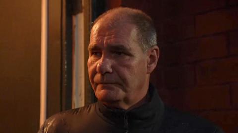 Steve, clean shaven with short grey hair, wearing a brown jacket zipped up standing outside a house 