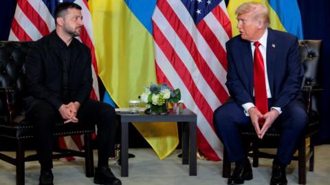 Zelensky wearing a black suit sits opposite Trump wearing a blue suit and red tie, with Ukraine and USA flags behind them