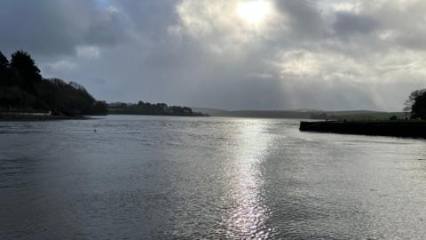 Kingsbridge estuary with the sun setting. It is a cloudy day and in the distance there is land either side of the estuary. The sun is coming through the clouds at the centre and is reflecting off the water.