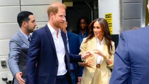 The Duke and Duchess of Suffolk walk out of a door while smiling. They are both wearing suits. Prince Harry is wearing a black blazer and trousers while Meghan is wearing a beige blazer and trousers. Both are wearing a white shirt. Prince Harry has ginger hair and blue eyes. Meghan has long brown hair and brown eyes.