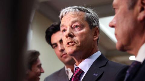 Andrew Bayly talks to reporters, wearing a suit and a purple tie.