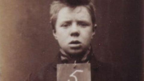 A mugshot of a young male prisoner., John Porter He is baby-faced with a mop of brown hair and is holding a sign with a 5 on it - the rest of the sign is cut off. The image is in sepia tones, and was taken on an old film camera. 