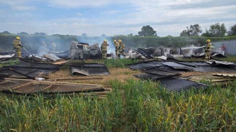 Seven firefighters in beige operational kit and yellow helmets spray water onto charred and twisted corrugated metal in a field. Smoke is still rising from the buildings' remains.
