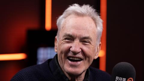 Larry Lamb, a man smiling at the camera. He is sitting next to a microphone.
He is wearing a black top. 