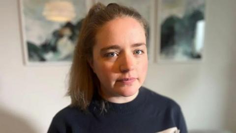 Stephanie Flynn faces the camera, wearing a navy jumper with her brown hair tied up in a high ponytail.
