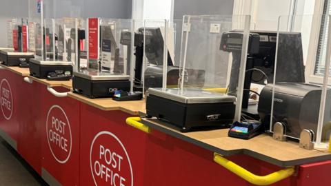 A Post Office counter with weighing machines, payment pads and computers. There is a perspex screen on the counter.