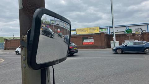 A convex mirror strapped to a lamppost on a traffic island at the junction of Lady Bay Bridge and Meadow Lane. Cars can be seen in the reflection of the mirror.