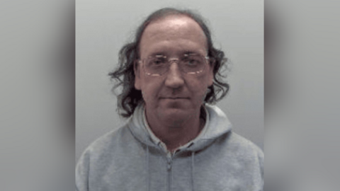 Police mugshot of a balding man with straggly long black hair and glasses. He is wearing a grey hoodie.   