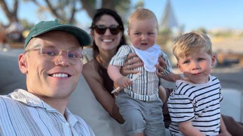 A man, woman and two young children. The man is wearing a green cap and glasses and the woman is holding one of the children, who is a baby.