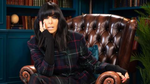 Claudia Winkleman sits on a brown, vintage-style, armchair, she is wearing a plaid suit, she rests her head on her left arm and stares into the camera. 