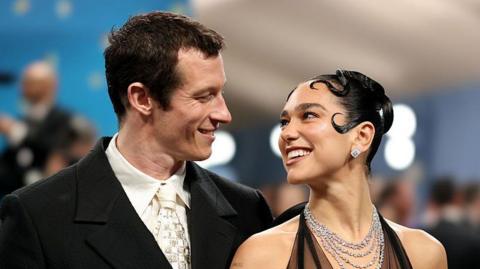 Dua Lipa and Callum Turner smile at each other - she wears a multi-stranded sparkling necklace and halterneck outfit, and has slicked back hair styled in curls around her face, while Turner wears a dark suit and cream tie and shirt 