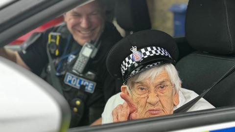 Gwen Lewis in police car looking through a window and pointing a finger 