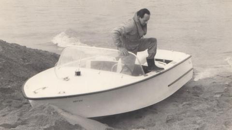 A black and white photograph of a speedboat docked on a sandbank on the edge of a large lake. Donald Campbell is stepping out of the boat on to the bank. 