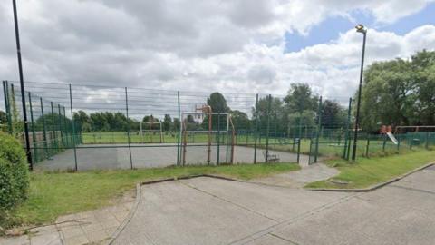 A play area surrounded by high green fences with football goals and basketball hoops at opposite ends. There is a paved area in the foreground and a playing field behind surrounded by trees. To the right is a playground area with a slide and other equipment.