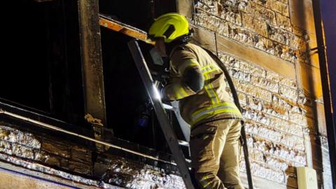 A firefighter on a ladder looks into the upper window of a building
