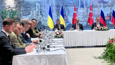 Row of men sat at long white table with microphones, Ukraine, Turkish and Russian flags behind them.