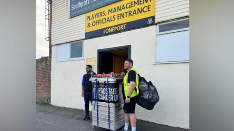 The pair are stood outside the entrance to Stockport FC with a large metal crate or luggage box. It has straps on it. Tom is wearing a high-vis jacket and has a large bag on his shoulder. Kyran is wearing dark clothing. Both men have beards.
