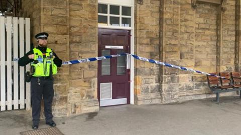 A police officer stands beside a stone building entrance that has been cordoned off with blue-and-white police tape. The taped‑off area includes a closed maroon door set into the building’s wall. Nearby, several wooden station benches sit along the platform area under a covered structure.
