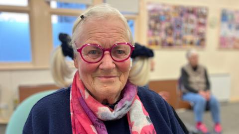 An elderly women is smiling at the camera. She has pink framed round glasses on and a pink and purple scarf. She has her hair in pigtails with a black scrunchies. 