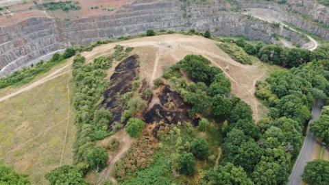Ariel view of the aftermath of the large grassland fire at Croft Hill, Leicestershire, showing the damaged area, which is surrounded by green trees and open space.