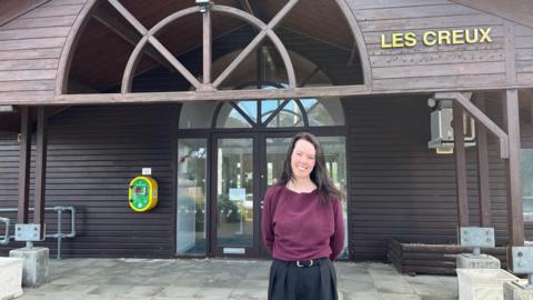 Jennifer Heald pictured outside Les Creux pavilion. She is wearing a plum coloured top and and is smiling. She has dark brown hair. In the background, the entrance to Les Creux is visible. The pavilion is mostly made of wood.