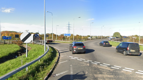 Cars are shown using a roundabout, with a grass verge to the left showing a national speed limit sign and a sign for the motorway ahead, with trees and an electricity pylon seen in the background.