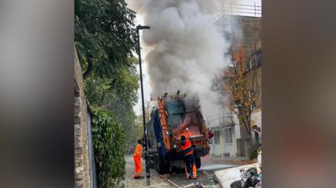 Refuse workers use hand held fire extinguishers to try and put the fire out in the back of a bin lorry. A large amount of smoke and some flames can be seen