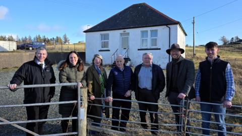The former nurses' cottage is in the background. It is a small single storey white washed building with steps leading up to the front door. A group of people involved in the project are standing in the foreground next to a fence.