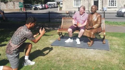 A man is photographing another man who sits on the bench next to Agatha Christie's statue in front of the Wallingford Museum with an open book. It's a sunny day.