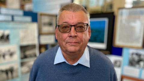 Adrian Maasz standing inside the RAF Marham Aviation Heritage Centre. He is looking directly at the camera and is wearing a blue and white patterned shirt with a blue jumper over the top and a pair of glasses.