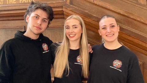 A teenage boy with medium-length brown hair and two teenage girls, one with long blonde hair and one with dark hair tied up in a ponytail. They're all wearing black tops branded with the Artemis College logo and are standing smiling, with their arms round each other, against a wood panelled wall.