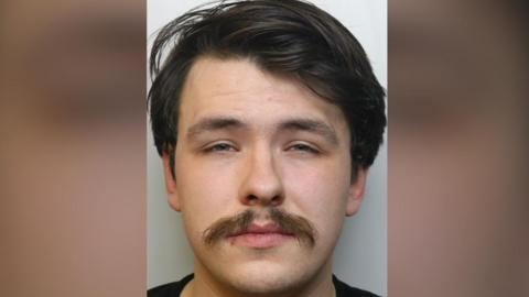 Close up image of young man with dark hair and bushy moustache wearing black t-shirt looking to camera