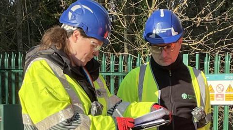 Two people in black Environment Agency fleece jumpers and yellow high-vis coats looking at a notepad. They also have blue Environment Agency hard hats on.